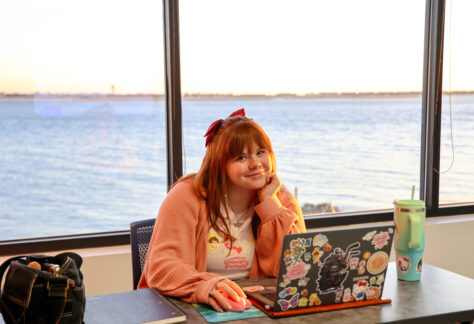 Young woman at a desk, smiling, with a laptop and waterfront view behind her.