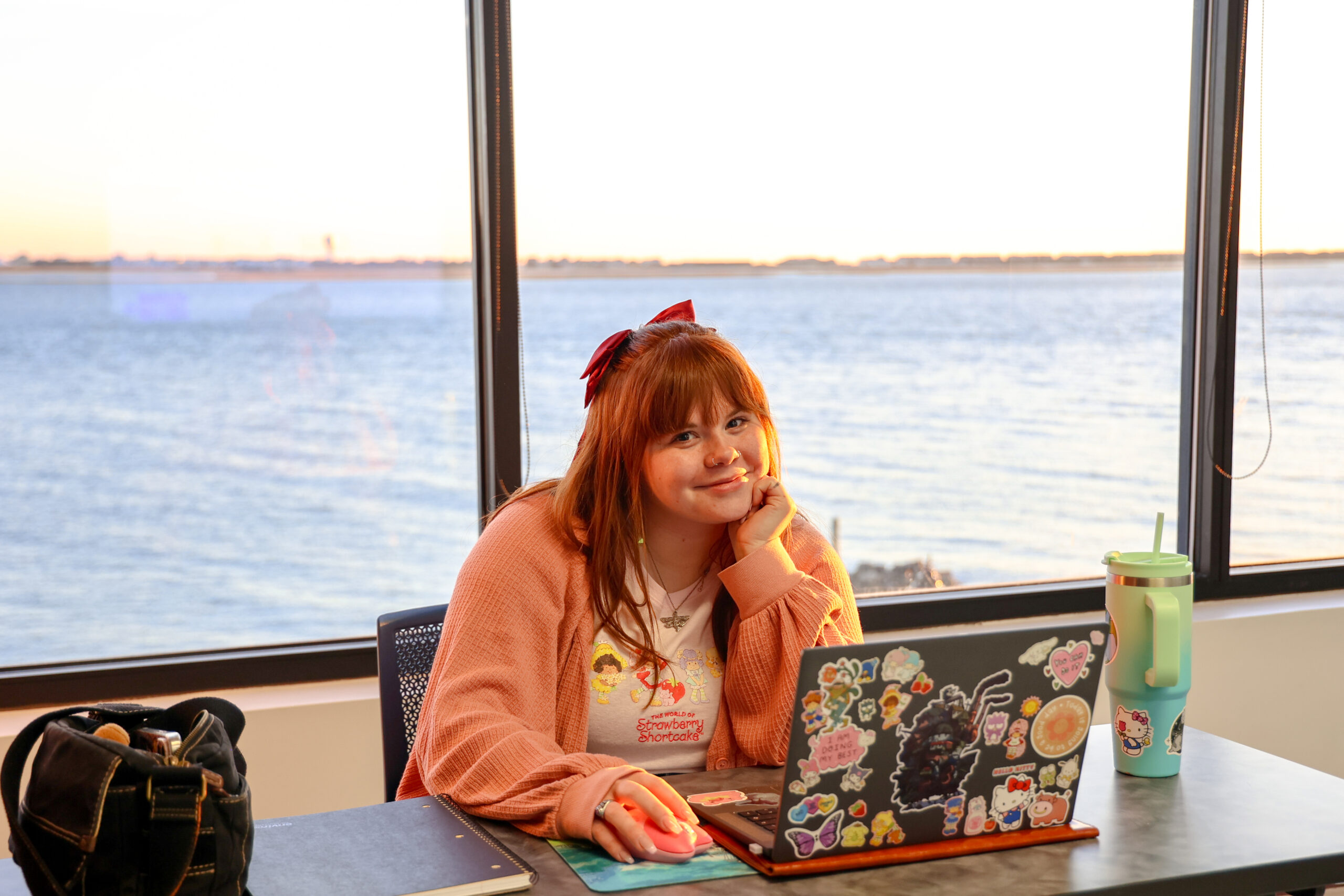 Young woman at a desk, smiling, with a laptop and waterfront view behind her.