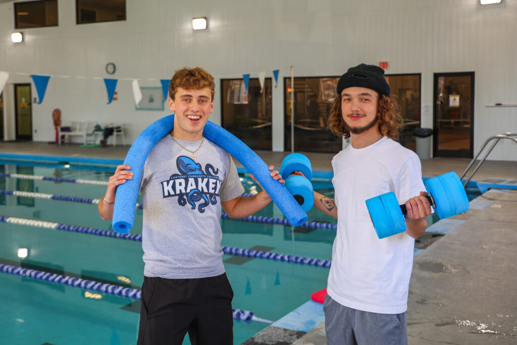 Two young men stand by an indoor pool, smiling at the camera. One wears a CarteretCC Kraken T-shirt with a blue pool noodle around his shoulders, while the other wears a white T-shirt and holds blue foam water dumbbells. The pool and lane dividers are visible in the background.