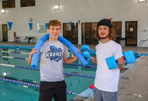 Two young men stand by an indoor pool, smiling at the camera. One wears a CarteretCC Kraken T-shirt with a blue pool noodle around his shoulders, while the other wears a white T-shirt and holds blue foam water dumbbells. The pool and lane dividers are visible in the background.