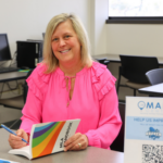 A smiling woman with blonde hair sits at a table in a classroom, wearing a bright pink blouse. She is holding a pen and an MLA handbook, appearing engaged in her work. The background features computers, chairs, and a window letting in natural light. A sign on the table reads "MAPS Center" with a QR code, encouraging students to provide feedback.