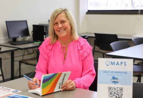 A smiling woman with blonde hair sits at a table in a classroom, wearing a bright pink blouse. She is holding a pen and an MLA handbook, appearing engaged in her work. The background features computers, chairs, and a window letting in natural light. A sign on the table reads "MAPS Center" with a QR code, encouraging students to provide feedback.