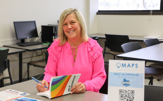 A smiling woman with blonde hair sits at a table in a classroom, wearing a bright pink blouse. She is holding a pen and an MLA handbook, appearing engaged in her work. The background features computers, chairs, and a window letting in natural light. A sign on the table reads "MAPS Center" with a QR code, encouraging students to provide feedback.