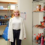 A student wearing glasses, a white sweater, and a white coat stands inside The Galley food pantry, holding a reusable shopping bag. Shelves stocked with snacks, beverages, canned goods, and hygiene products are visible in the background. A sign on the wall displays information about the pantry's services.