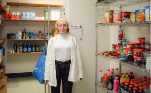 A student wearing glasses, a white sweater, and a white coat stands inside The Galley food pantry, holding a reusable shopping bag. Shelves stocked with snacks, beverages, canned goods, and hygiene products are visible in the background. A sign on the wall displays information about the pantry's services.