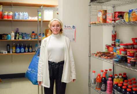 A student wearing glasses, a white sweater, and a white coat stands inside The Galley food pantry, holding a reusable shopping bag. Shelves stocked with snacks, beverages, canned goods, and hygiene products are visible in the background. A sign on the wall displays information about the pantry's services.