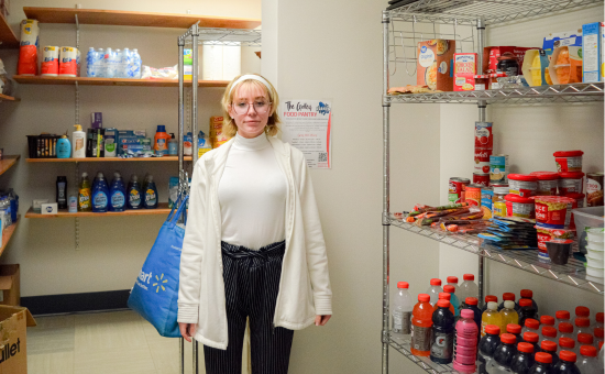 A student wearing glasses, a white sweater, and a white coat stands inside The Galley food pantry, holding a reusable shopping bag. Shelves stocked with snacks, beverages, canned goods, and hygiene products are visible in the background. A sign on the wall displays information about the pantry's services.