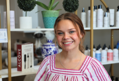 Young woman with light brown hair tied back, wearing a white blouse with pink stripes, smiling warmly in a salon setting. She is standing in front of shelves stocked with haircare products and decorative items.