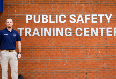 A man wearing a navy blue shirt and khaki pants stands in front of a brick wall with large white letters that read "Public Safety Training Center."