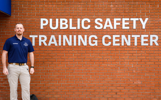A man wearing a navy blue shirt and khaki pants stands in front of a brick wall with large white letters that read "Public Safety Training Center."
