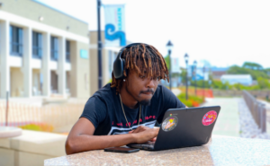 Student with headphones working on a sticker-covered laptop at an outdoor campus table.