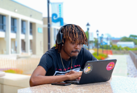 Student with headphones working on a sticker-covered laptop at an outdoor campus table.