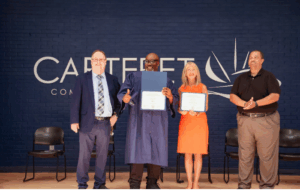 A graduate wearing a navy blue cap and gown stands proudly at the center of a stage holding a diploma, smiling and giving a thumbs-up. He is joined by three individuals: a man in a suit and tie on the left, a woman in an orange dress holding a certificate, and a man in a black polo shirt applauding on the right. Behind them is a dark blue brick wall with the Carteret Community College logo. Four empty chairs are visible in the background.