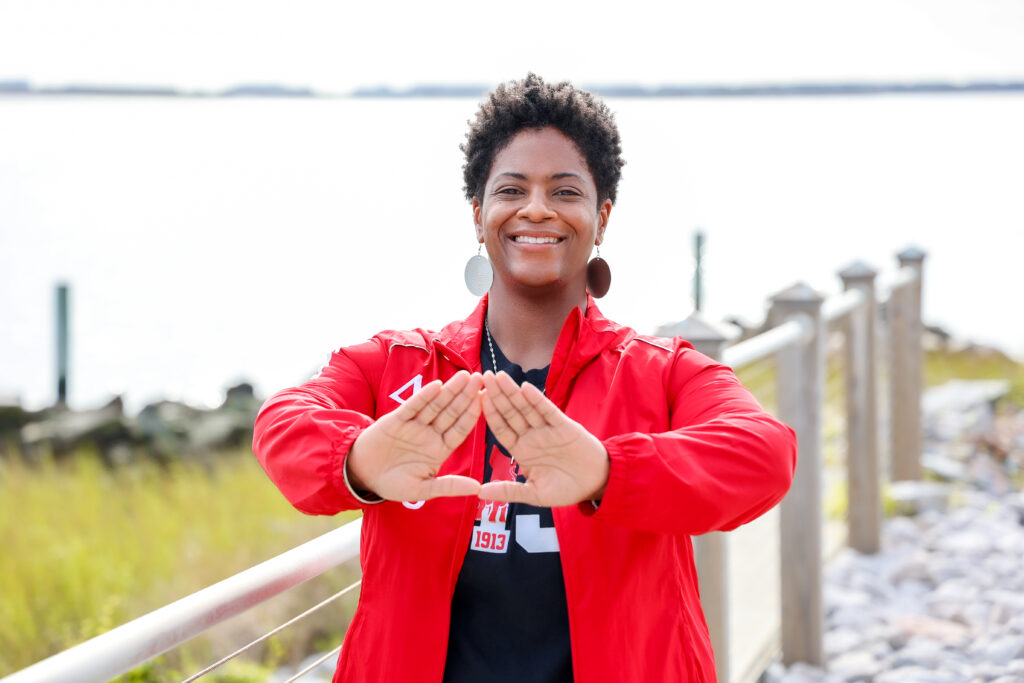 A smiling woman stands outdoors near a waterfront railing, wearing a red jacket and large round earrings. She holds her hands up in front of her forming a triangle shape. The background shows calm water, greenery, and a bright sky.