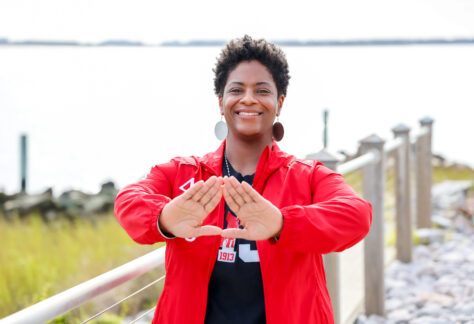 A smiling woman stands outdoors near a waterfront railing, wearing a red jacket and large round earrings. She holds her hands up in front of her forming a triangle shape. The background shows calm water, greenery, and a bright sky.