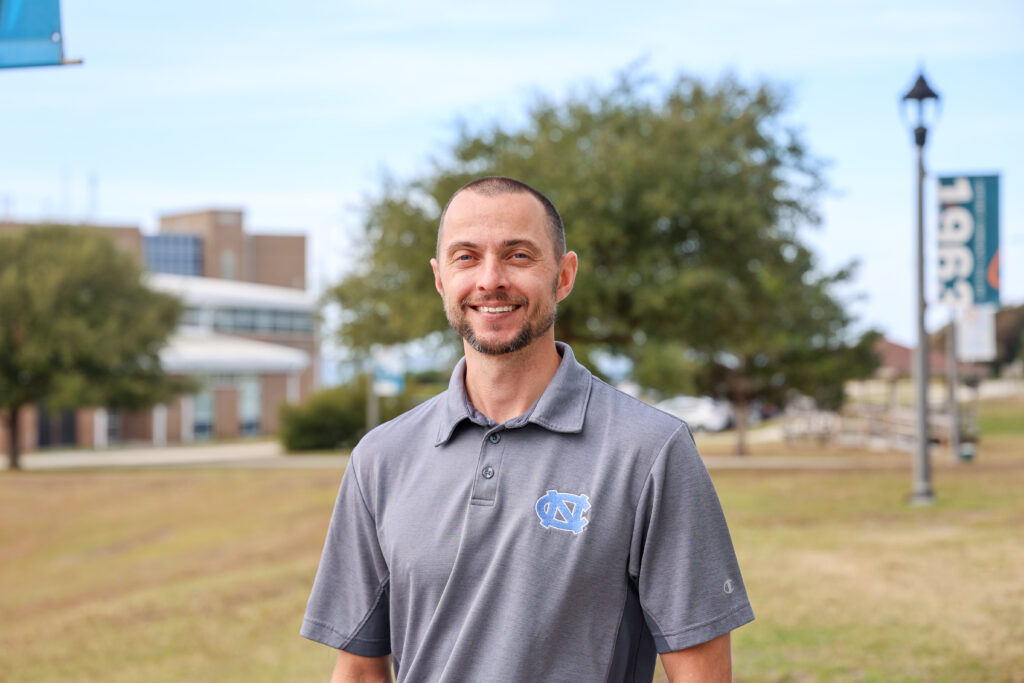a male standing outside on campus wearing a grey shirt with the UNC logo