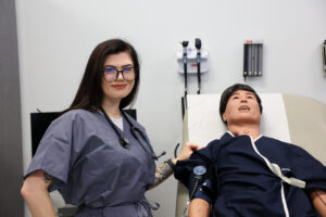 Geneveive Muehlenkamp, a Carteret Community College Medical Assisting student, smiles while standing beside a medical training mannequin in a clinical lab setting. She’s wearing gray scrubs and a stethoscope, demonstrating confidence and professionalism as she practices her clinical skills.