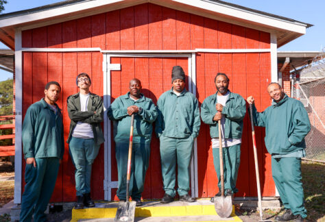 A group of six men wearing green work uniforms stand in front of a bright red shed on a sunny day. Some hold shovels and tools, suggesting participation in an outdoor training or vocational program. They stand side by side, smiling or posing confidently, with a fenced area and buildings visible in the background.