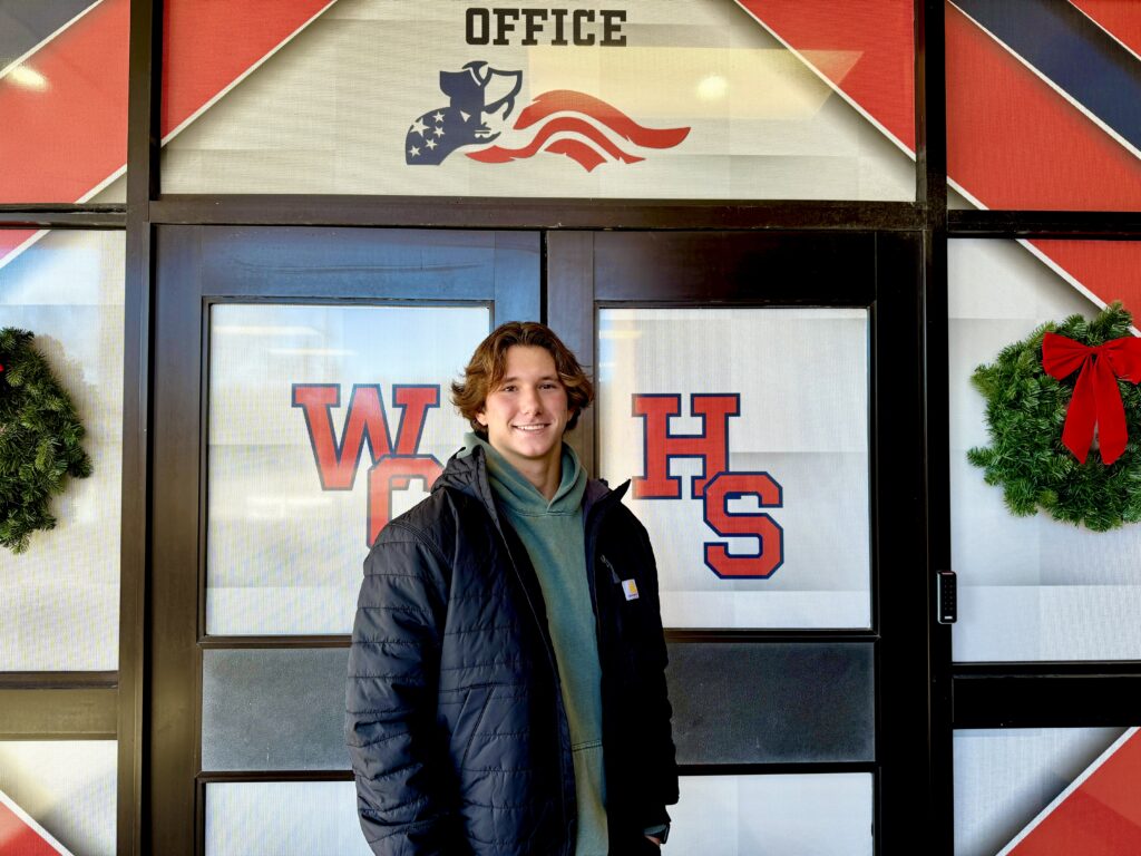A male student standing outside of West Carteret high school doors with the logo showing