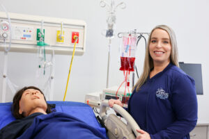 Professional photo of a woman wearing a navy Carteret Community College Associate Degree Nursing jacket, standing beside a hospital bed in a clinical simulation lab. A medical training mannequin lies in the bed, with IV bags, oxygen outlets, and monitoring equipment visible in the background.