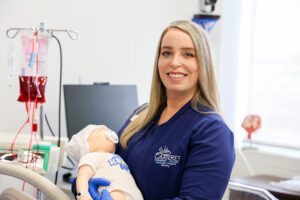 Professional photo of a woman wearing a navy jacket embroidered with the Carteret Community College Associate Degree Nursing logo, smiling while holding a medical training mannequin in a clinical lab setting. An IV bag filled with red fluid and medical equipment are visible in the background.