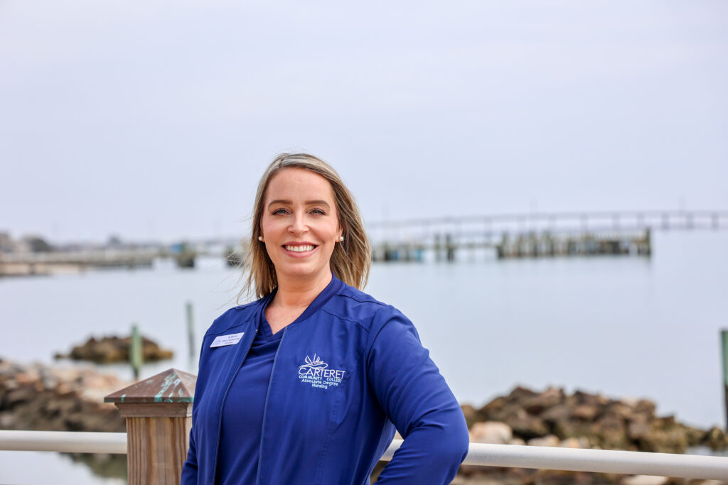 Professional headshot of a smiling woman wearing a navy jacket embroidered with the Carteret Community College Associate Degree Nursing logo, standing outdoors by the water with a dock and rocky shoreline softly blurred in the background.