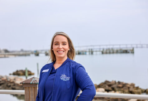 Professional headshot of a smiling woman wearing a navy jacket embroidered with the Carteret Community College Associate Degree Nursing logo, standing outdoors by the water with a dock and rocky shoreline softly blurred in the background.