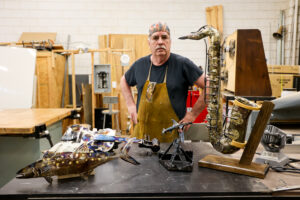 Man stands behind a worktable displaying welded metal artwork including a fish, violin, saxophone, and small sculptures in a workshop.