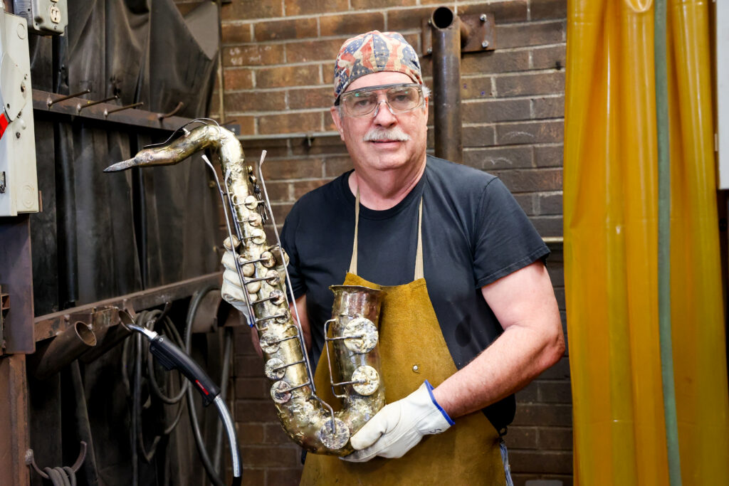 Man in a welding shop wearing safety glasses and an apron holds a handcrafted metal saxophone sculpture.