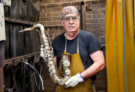 Man in a welding shop wearing safety glasses and an apron holds a handcrafted metal saxophone sculpture.