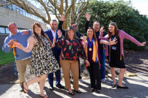 A group of people stand outdoors on a sunny day smiling and posing energetically with raised hands, with trees and a campus building in the background.