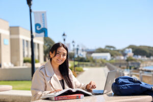 A woman sits outdoors at a waterfront table studying with an open textbook and laptop, with a backpack beside her and a sunny coastal walkway in the background.