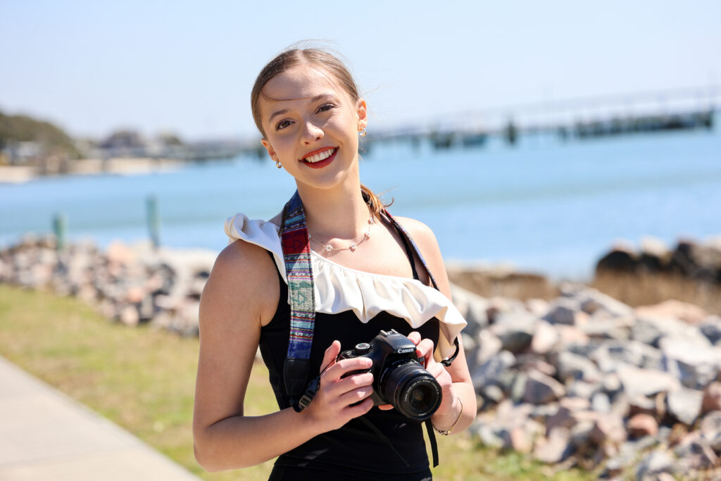 A smiling woman stands outdoors near the water holding a DSLR camera, with a rocky shoreline and bridge in the background on a sunny day.