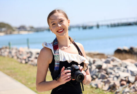 A smiling woman stands outdoors near the water holding a DSLR camera, with a rocky shoreline and bridge in the background on a sunny day.