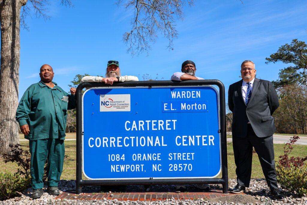 Four men stand behind a Carteret Correctional Center sign in Newport, North Carolina, representing a partnership between the correctional facility and Carteret Community College.