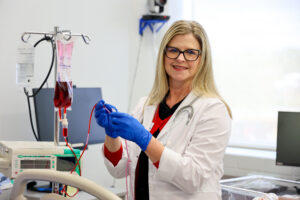 A healthcare professional wearing a lab coat and blue gloves stands in a clinical setting beside medical equipment, including an IV bag filled with red fluid, while smiling and preparing for a procedure.
