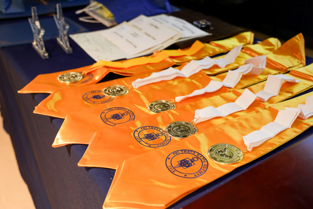 Phi Theta Kappa honor cords with gold medallions and white ribbons neatly arranged on a table alongside induction certificates, prepared for a college honor society ceremony.