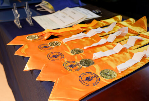 Phi Theta Kappa honor cords with gold medallions and white ribbons neatly arranged on a table alongside induction certificates, prepared for a college honor society ceremony.