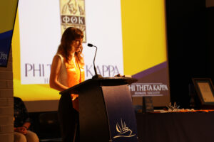 BV4A0286 Student wearing a Phi Theta Kappa stole speaks at a podium during an induction ceremony, with the PTK logo displayed on a screen behind her.