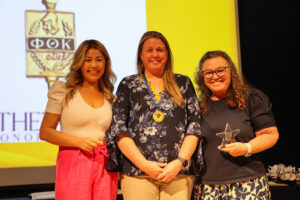 BV4A0338 Three women smile while holding recognition awards and wearing Phi Theta Kappa medals on stage during the ceremony.