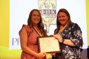 BV4A0484 Two women hold a framed Phi Theta Kappa certificate while smiling on stage during the induction ceremony.