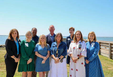 A group of nine adults stand together outdoors near the water, smiling at the camera, with a clear sky and shoreline in the background.