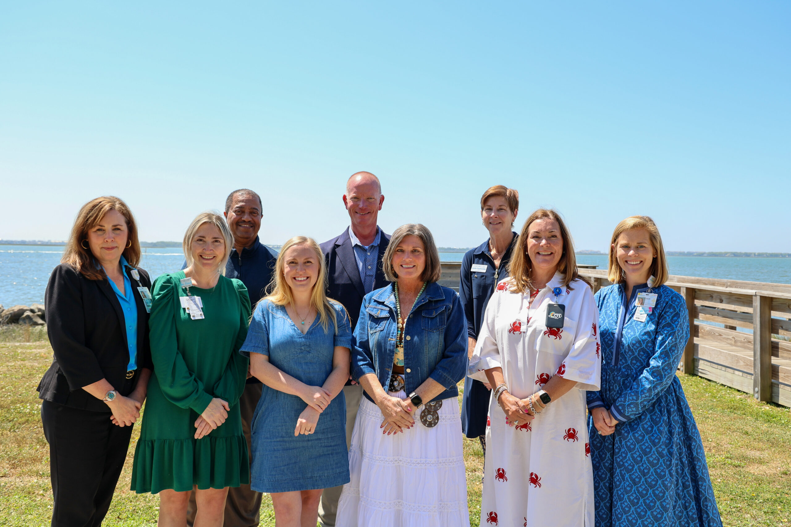 A group of nine adults stand together outdoors near the water, smiling at the camera, with a clear sky and shoreline in the background.