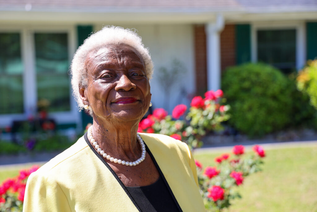 Portrait of Barbara Horton Hill standing outdoors in front of a home and garden, wearing a yellow blazer and pearl necklace, smiling.