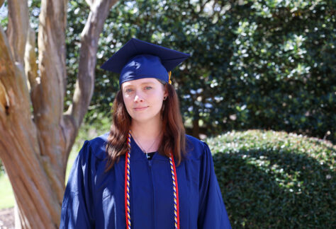 Female student in a blue cap and gown standing outside.