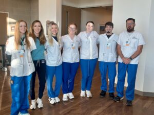 5 women and 2 men in hospital attire standing in the hallway smiling